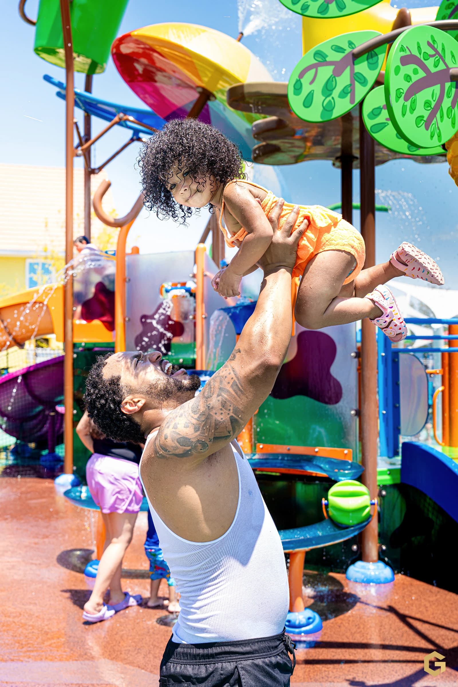 Edgardo joyfully lifts his daughter above the splash pad