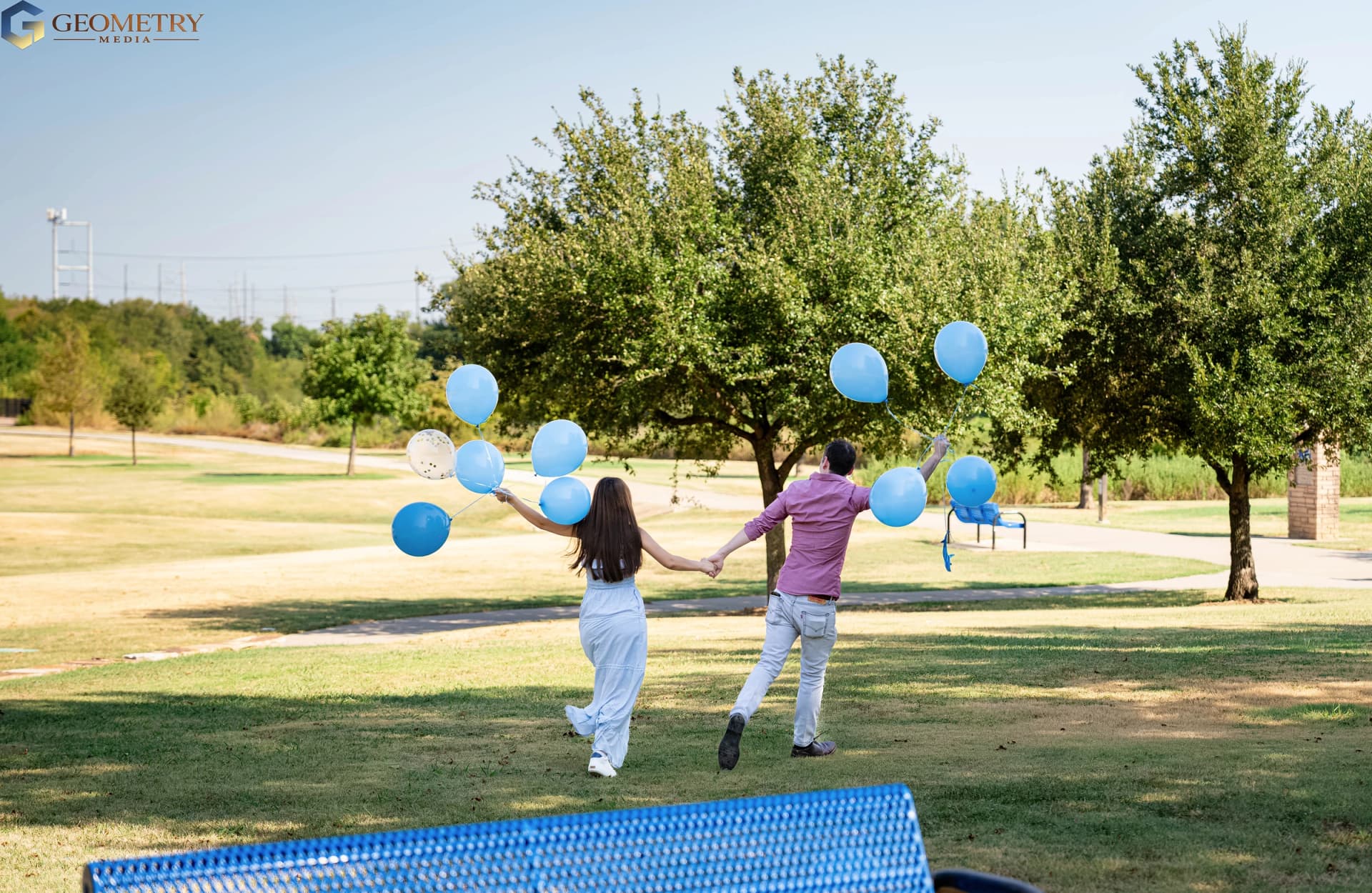 The couple walk hand in hand across the lawn carrying blue balloons