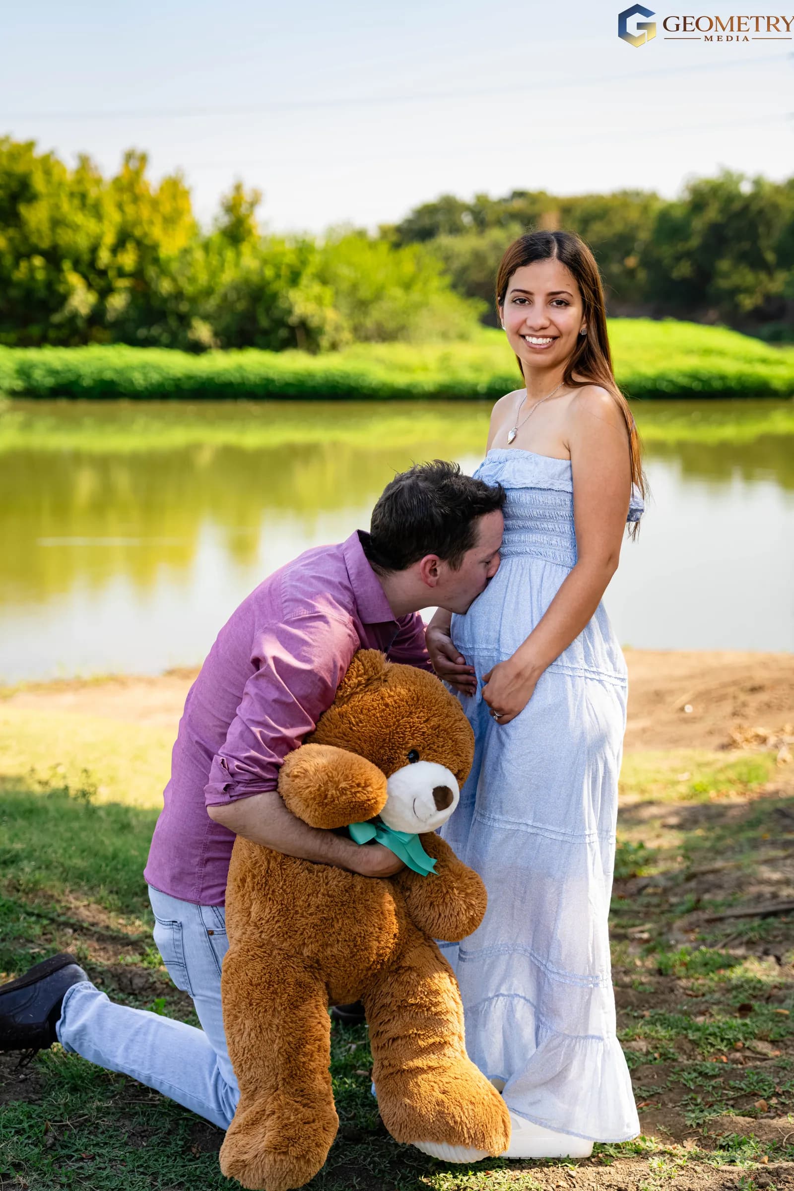Expectant parents pose beside the water with a large teddy bear