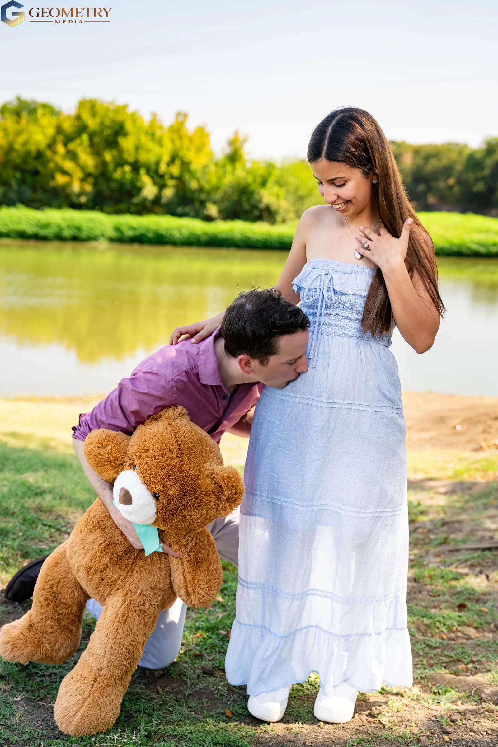 Mother smiles while the father kisses her baby bump beside the lake